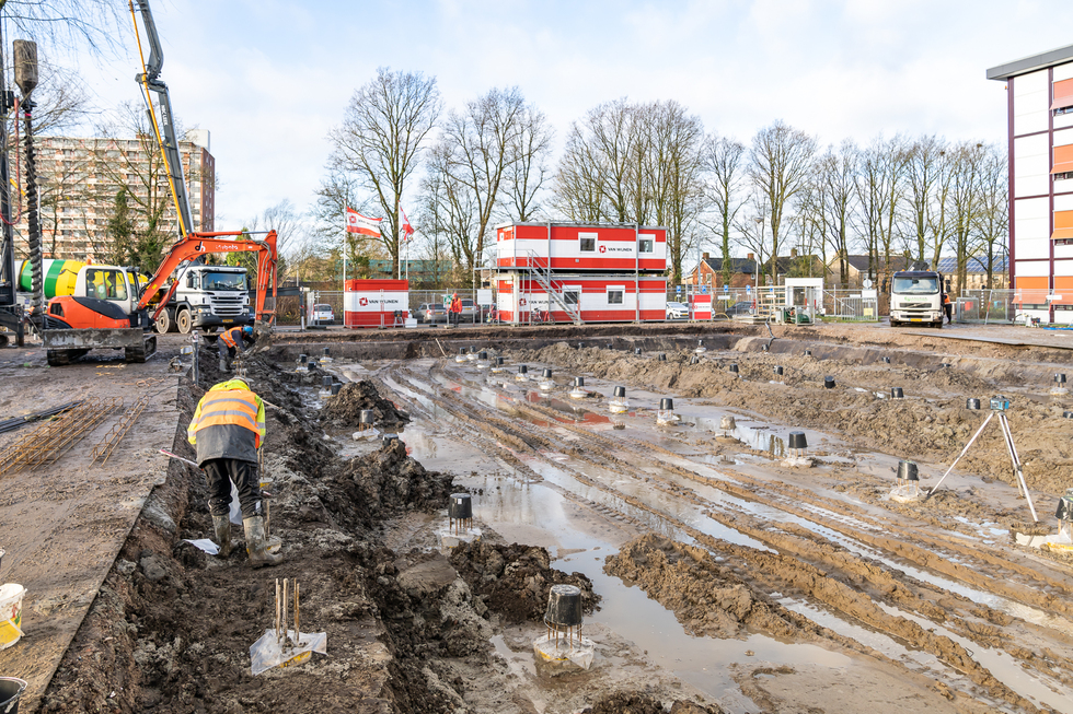 Bauarbeiter bei Fundamentarbeiten auf schlammiger Baustelle mit Baumaschinen und Absperrungen im Hintergrund