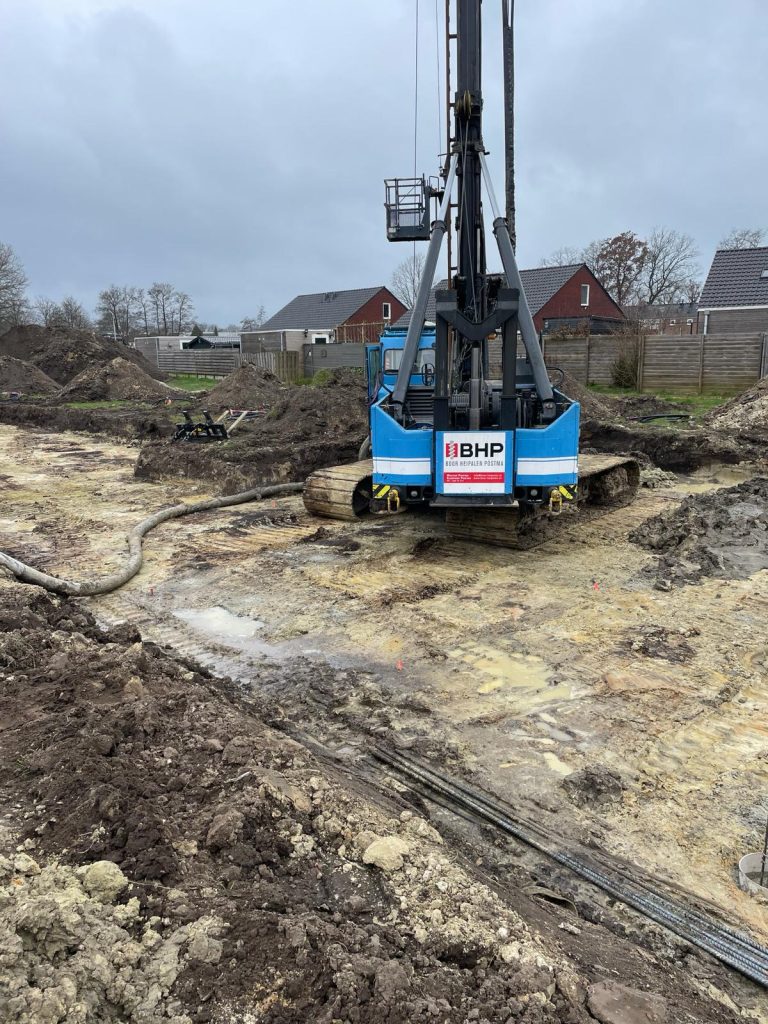Rammgerät auf Raupenfahrwerk bei Pfahlgründungsarbeiten auf schlammiger Baustelle mit Wohnhäusern im Hintergrund